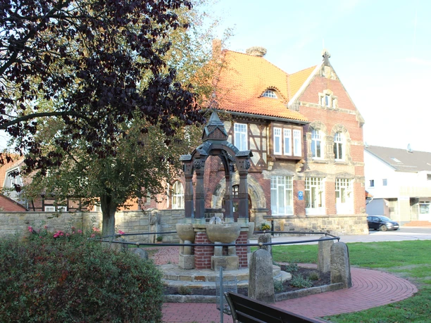 Historischer Brunnen in Rehburgs Ortsmitte, flankiert von Bäumen und einem stattlichen Fachwerkhaus.