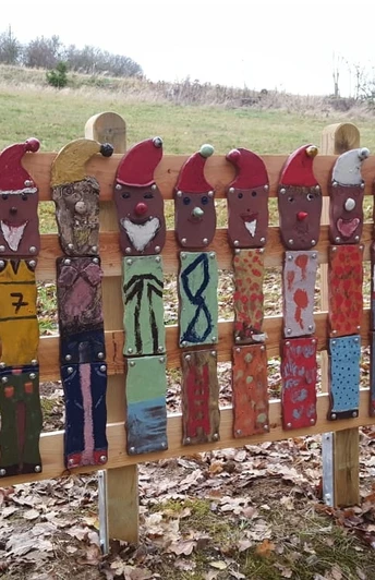 Zwölf bemalte Holzlatten in Zwergenform stehen auf einem Laubboden in einer ländlichen Landschaft.