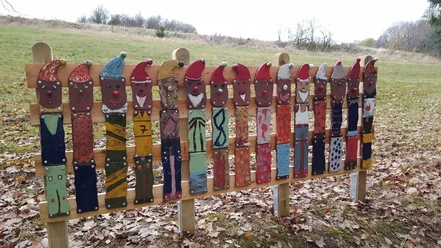 Skulpturen am Wegesrand Bleiwäsche Zwölf bemalte Holzlatten in Zwergenform stehen auf einem Laubboden in einer ländlichen Landschaft.