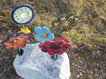 Bunte Keramikblumen, kunstvoll gestaltet, stehen auf einem Stein inmitten von Gras und Laub.