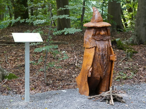 Brüder Grimm Märchenwanderweg Bad Rehburg Holzskulptur eines stehenden, bärtigen Zauberers mit spitzem Hut, umgeben von Waldbäumen und Laub.