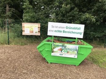 Grüner Container auf freier Fläche mit Schild zur Förderung der Kompostierung, umgeben von Bäumen.