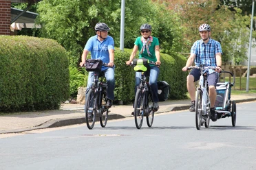 Three cyclists and a child in a bicycle trailer ride relaxed along a residential street.