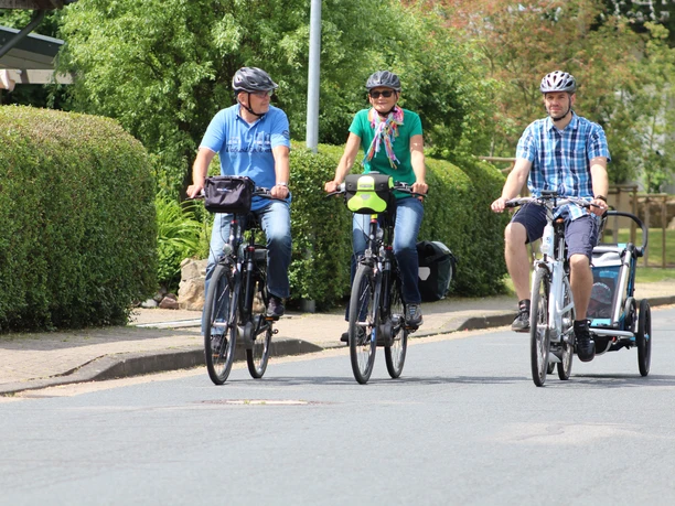 Three cyclists and a child in a bicycle trailer ride relaxed along a residential street.