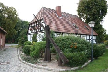 Ein historischer Ziehbrunnen steht vor einem Fachwerkhaus umgeben von Büschen und bunten Blumen.