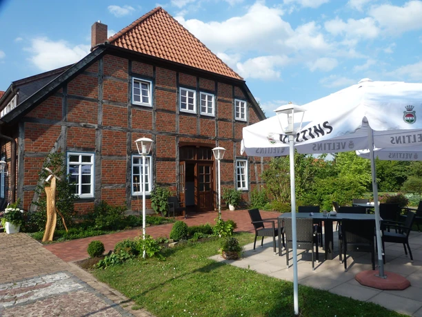 A brick house with a red roof and wooden frame, surrounded by plants and a parasol.