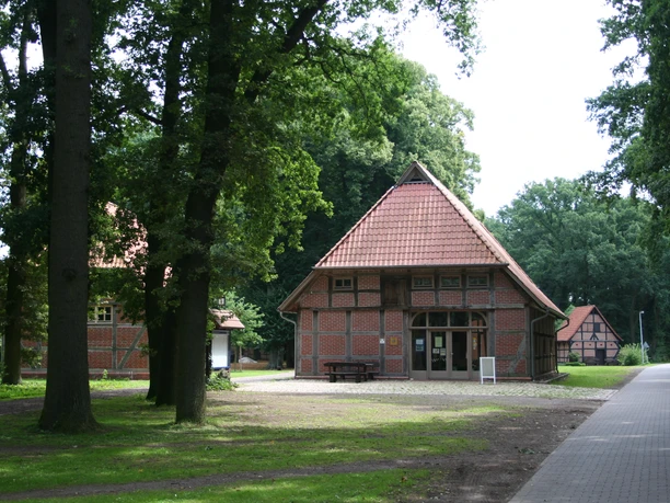 Brick building with gabled roof and half-timbered elements, surrounded by tall trees and forest path.