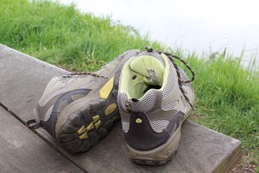 Two worn hiking boots stand on a wooden bench in front of a green meadow and a body of water.