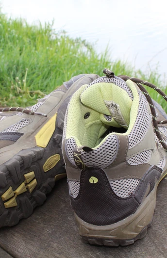 Zwei getragene Wanderschuhe stehen auf einer Holzbank vor einer grünen Wiese und einem Gewässer.