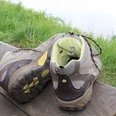 Wanderschuhe Zwei getragene Wanderschuhe stehen auf einer Holzbank vor einer grünen Wiese und einem Gewässer.