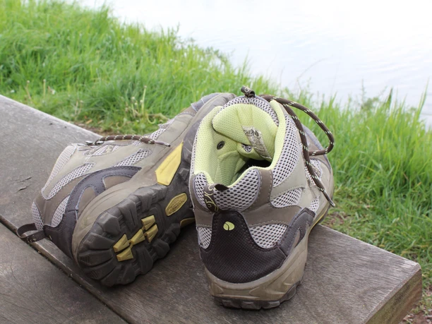 Wanderschuhe Zwei getragene Wanderschuhe stehen auf einer Holzbank vor einer grünen Wiese und einem Gewässer.