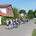 Wandertour "Op'n Land" (Auf dem Land) Eine Gruppe von Wanderern mit Rucksäcken spaziert auf einer Dorfstraße entlang traditioneller Häuser.