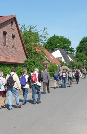 Wandertour "Op'n Land" (Auf dem Land) Eine Gruppe von Wanderern mit Rucksäcken spaziert auf einer Dorfstraße entlang traditioneller Häuser.