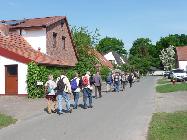 Wandertour "Op'n Land" (Auf dem Land) Eine Gruppe von Wanderern mit Rucksäcken spaziert auf einer Dorfstraße entlang traditioneller Häuser.