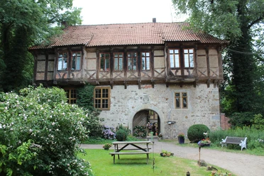 Rittergut Brokeloh Ein historisches Fachwerkhaus mit rotem Ziegeldach, umgeben von grünen Bäumen und Blumenbeeten.