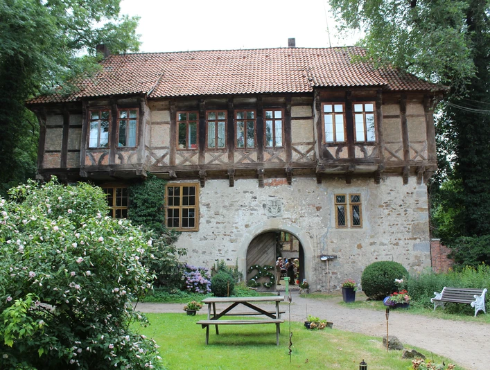 Rittergut Brokeloh Ein historisches Fachwerkhaus mit rotem Ziegeldach, umgeben von grünen Bäumen und Blumenbeeten.