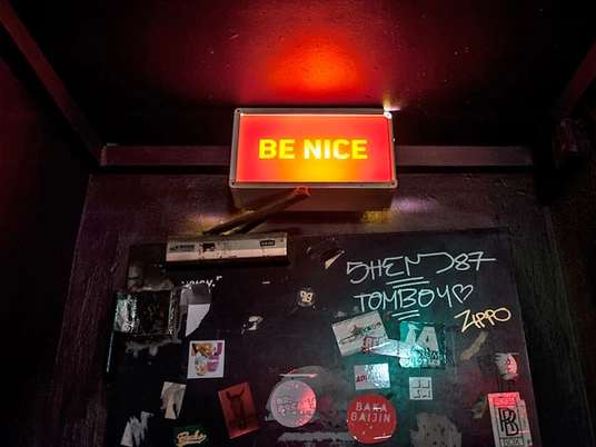 Subway Das Bild zeigt eine dunkle Wand mit einem leuchtend roten Schild, auf dem "BE NICE" steht. Darunter sind Graffiti und bunte Sticker zu sehen.The picture shows a dark wall with a bright red sign that reads "BE NICE". Graffiti and colorful stickers can be seen underneath.
