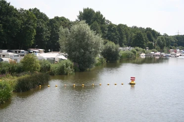 Campingplatz und Yachtclub Lahde Ruhiger See mit Segelbooten und Wohnmobilen am Ufer, umgeben von üppigen grünen Bäumen und Schilf.
