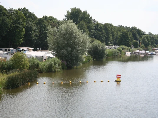 Campingplatz und Yachtclub Lahde Ruhiger See mit Segelbooten und Wohnmobilen am Ufer, umgeben von üppigen grünen Bäumen und Schilf.