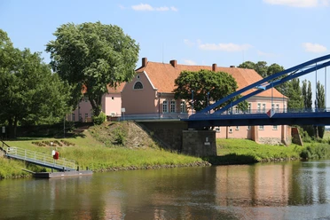 Hoya Ein rosa Fachwerkhaus an einem Flussufer, flankiert von großen Bäumen und einer blauen Brücke.
