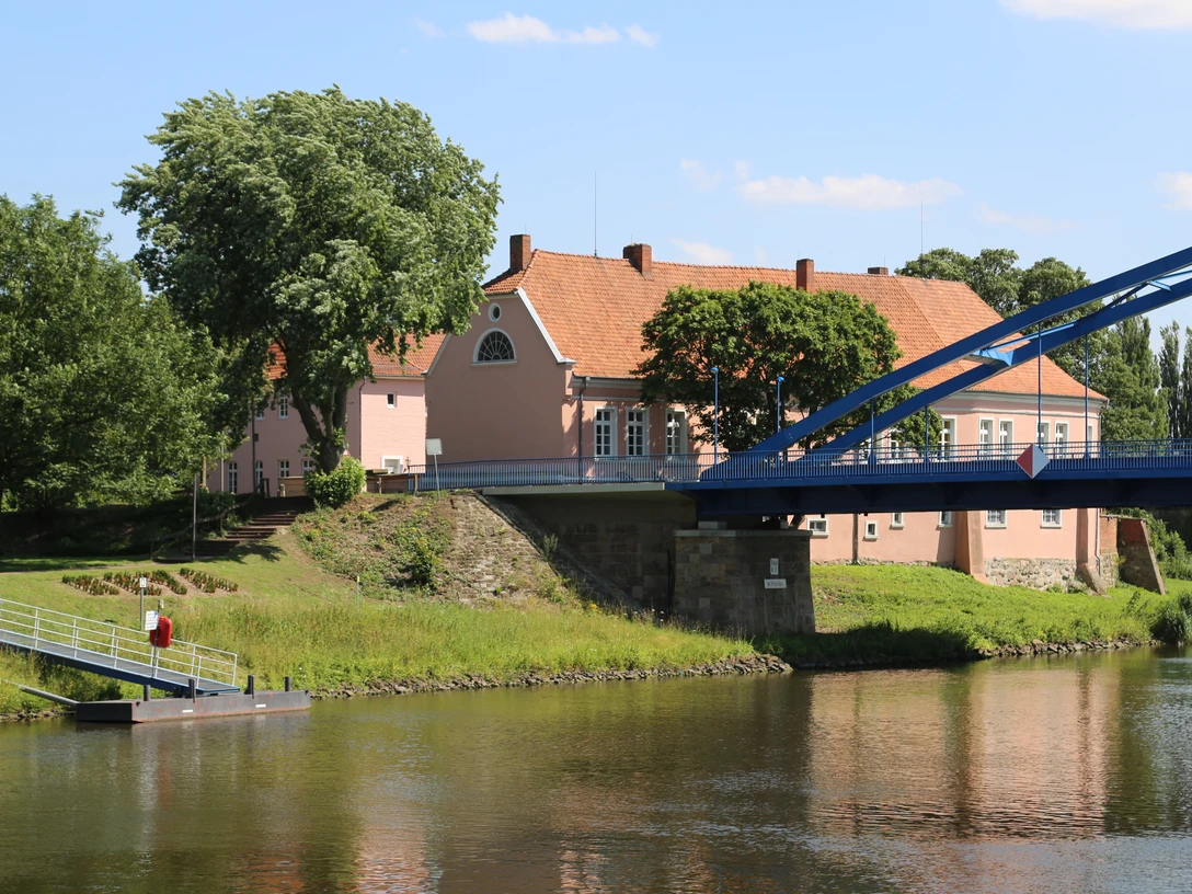 Hoya Ein rosa Fachwerkhaus an einem Flussufer, flankiert von großen Bäumen und einer blauen Brücke.
