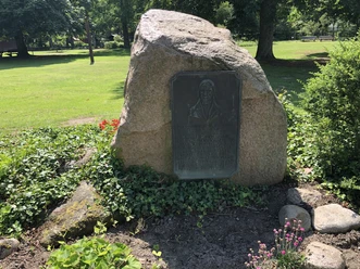 Gedenkstein mit Inschrift und Relief in einem ruhigen Park, von Pflanzen und Bäumen umgeben.