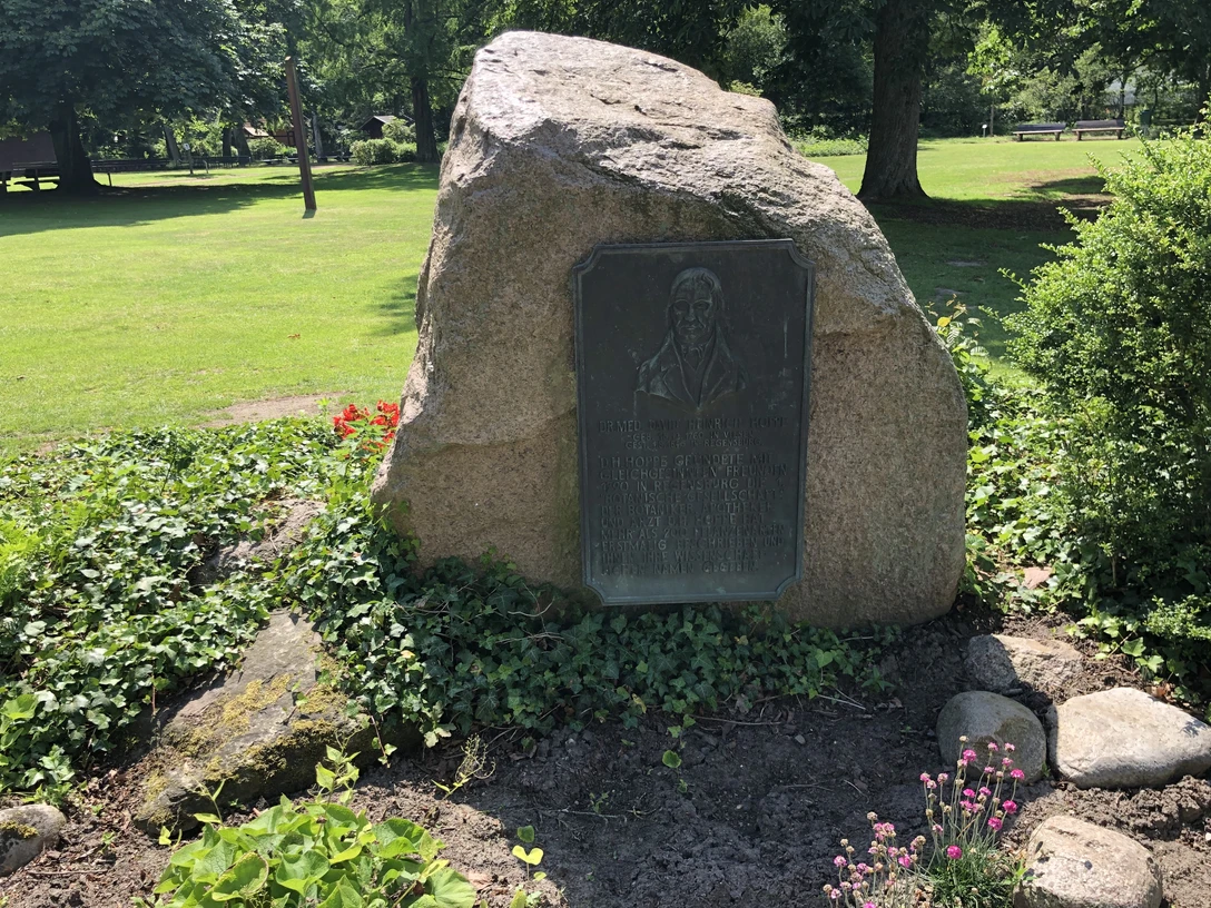 Gedenkstein mit Inschrift und Relief in einem ruhigen Park, von Pflanzen und Bäumen umgeben.