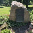 Gedenkstein mit Inschrift und Relief in einem ruhigen Park, von Pflanzen und Bäumen umgeben.
