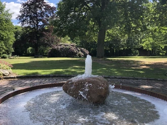 A rippling fountain stands in the center of a green park with lush trees and flowerbeds.