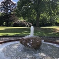 A rippling fountain stands in the center of a green park with lush trees and flowerbeds.