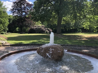 Kurpark Ein plätschernder Brunnen steht im Zentrum eines grünen Parks mit üppigen Bäumen und Blumenbeeten.