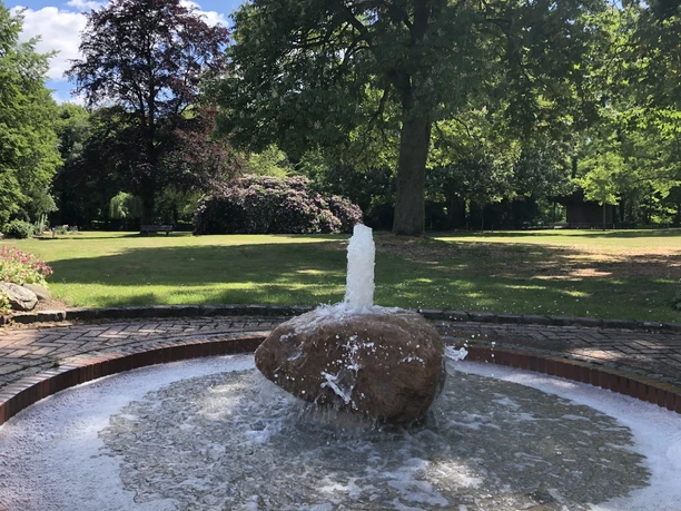 Kurpark Ein plätschernder Brunnen steht im Zentrum eines grünen Parks mit üppigen Bäumen und Blumenbeeten.