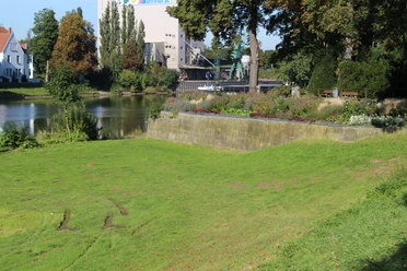 Grüne Wiese vor einem historischen Mauerabschnitt an einem Fluss, im Hintergrund Bäume und Gebäude.