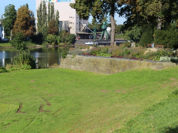 Grüne Wiese vor einem historischen Mauerabschnitt an einem Fluss, im Hintergrund Bäume und Gebäude.