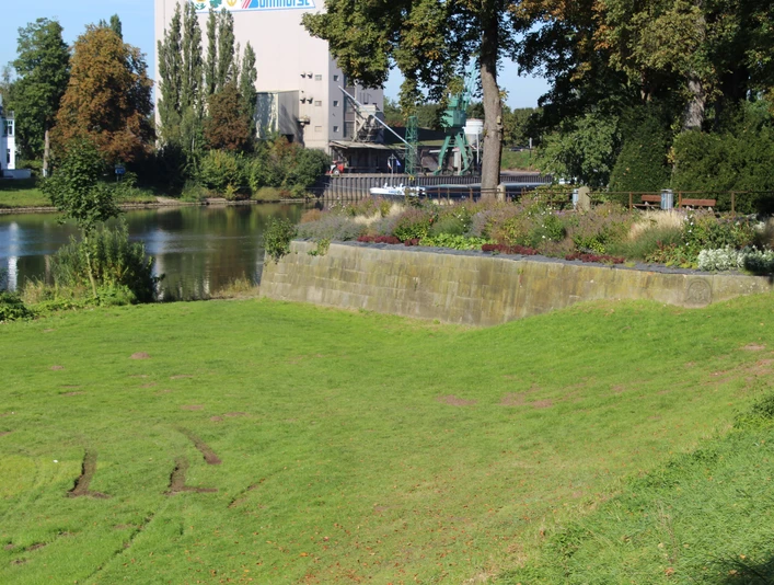 Grüne Wiese vor einem historischen Mauerabschnitt an einem Fluss, im Hintergrund Bäume und Gebäude.