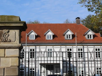 Fachwerkhaus mit rotem Ziegeldach und markantem Giebeldach, hinter schmiedeeisernem Zaun.