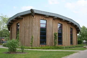 Ein modernes rundes Gebäude mit Holzfassade und markant geneigtem Metalldach, von Natur umgeben.