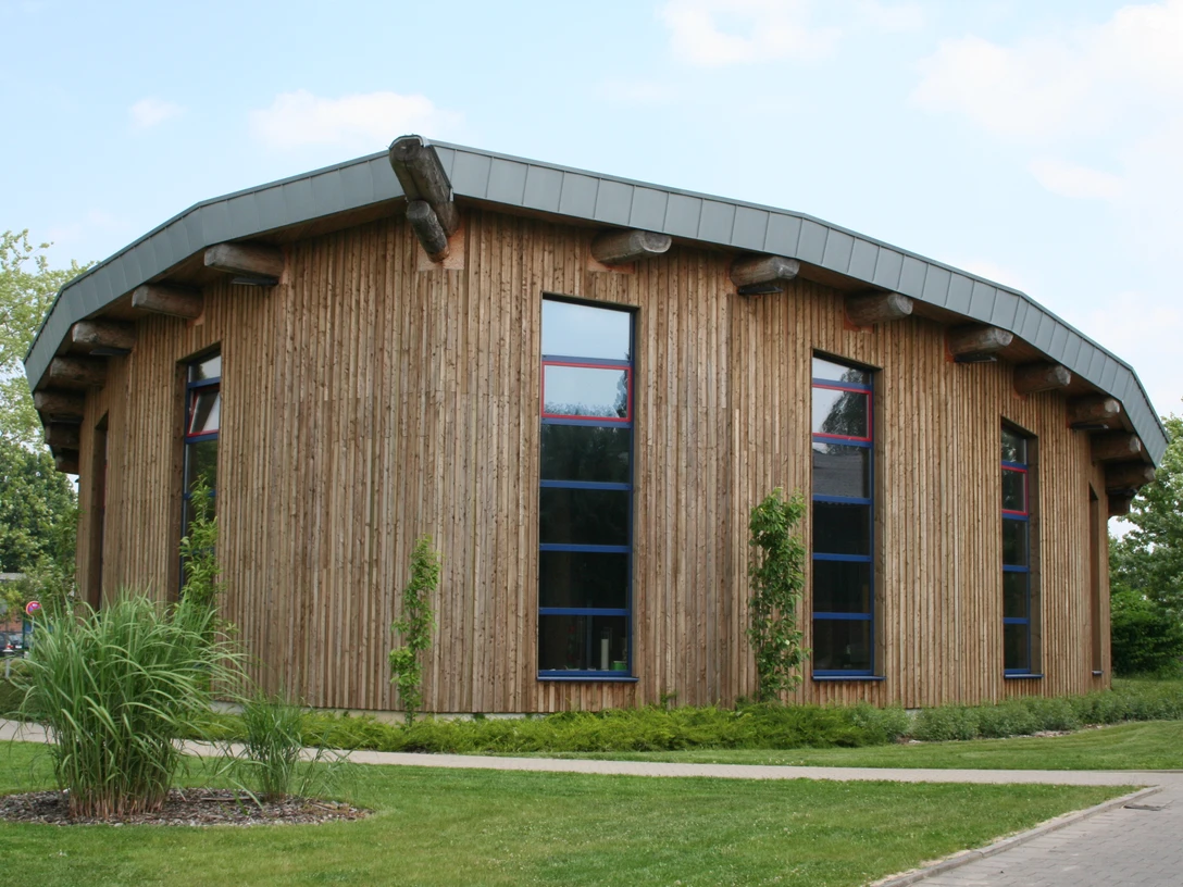 Deula Ein modernes rundes Gebäude mit Holzfassade und markant geneigtem Metalldach, von Natur umgeben.
