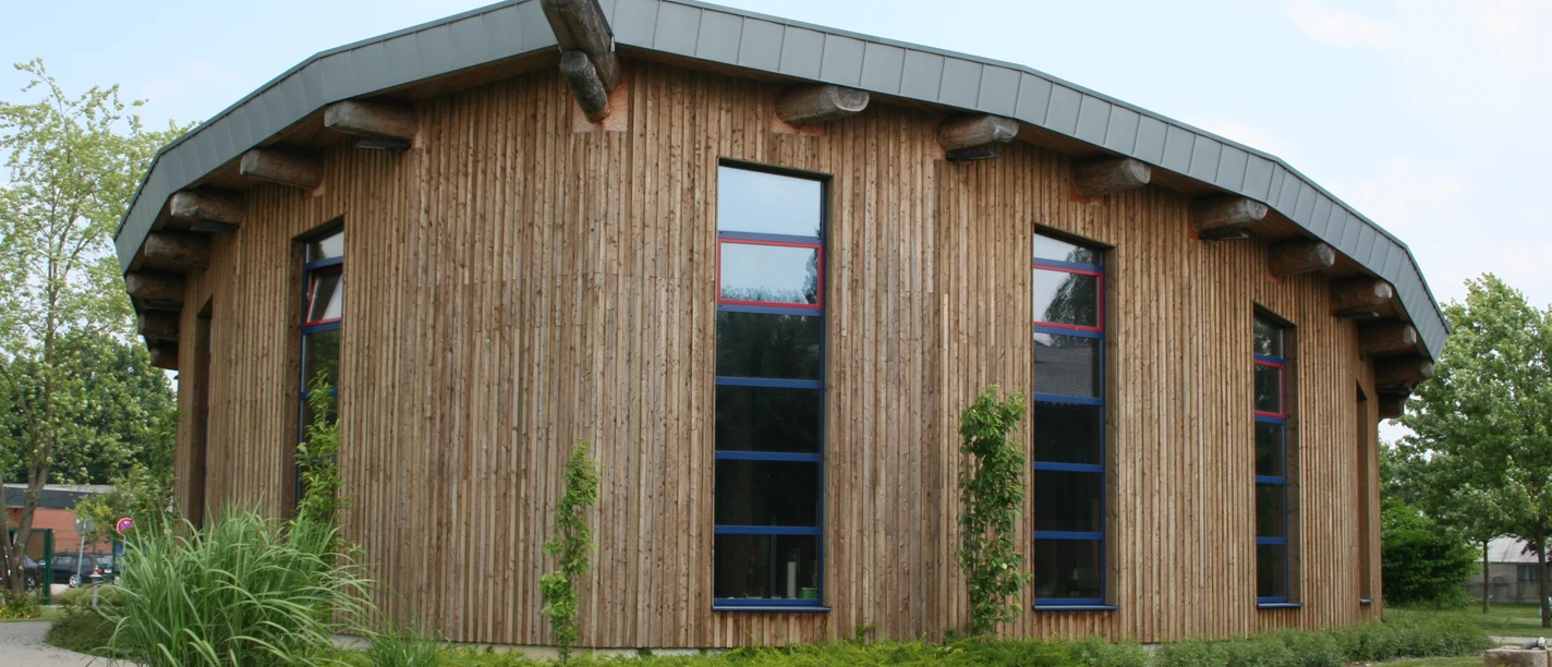Deula Ein modernes rundes Gebäude mit Holzfassade und markant geneigtem Metalldach, von Natur umgeben.