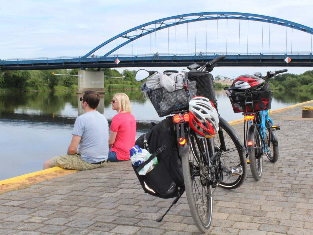 Zwei Fahrräder stehen am Flussufer; dahinter sitzen zwei Personen und blicken auf eine blaue Brücke.