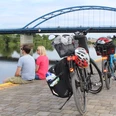 Zwei Fahrräder stehen am Flussufer; dahinter sitzen zwei Personen und blicken auf eine blaue Brücke.