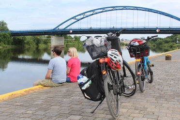 Radfahrer Zwei Fahrräder stehen am Flussufer; dahinter sitzen zwei Personen und blicken auf eine blaue Brücke.