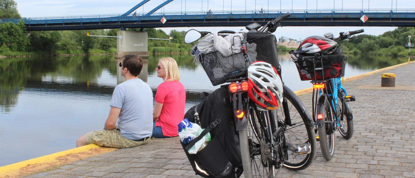 Radfahrer Zwei Fahrräder stehen am Flussufer; dahinter sitzen zwei Personen und blicken auf eine blaue Brücke.