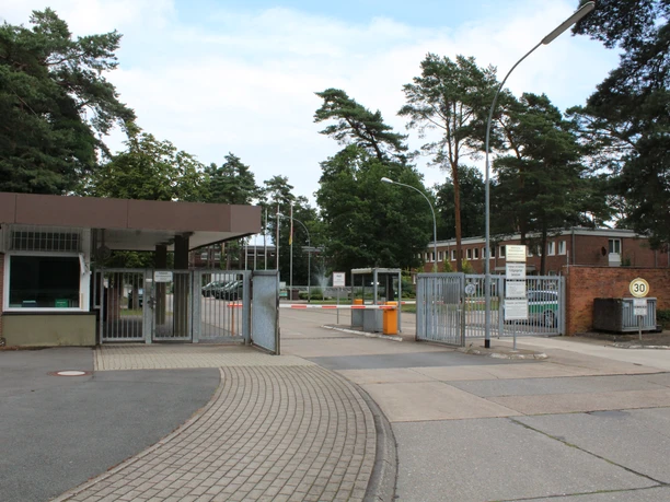 Eingang der Kaserne Langendamm, gesäumt von Kiefern und Backsteingebäuden, mit Schranken und Schildern.