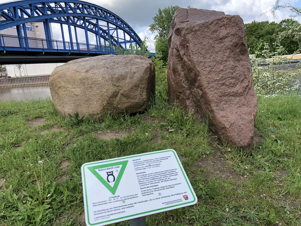 Zwei große Findlinge stehen auf einer Wiese, im Hintergrund ist eine blaue Brücke zu sehen.