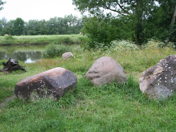 Wesersteine (3).JPG Drei große, bemooste Steine liegen in einer grünen Wiese vor einem Fluss, umgeben von Bäumen.