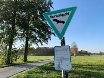 Naturschutzgebiet auf dem Ströhen Schild "Naturschutzgebiet" mit Adler, flankiert von Bäumen, vor einer weiten Wiesenlandschaft.