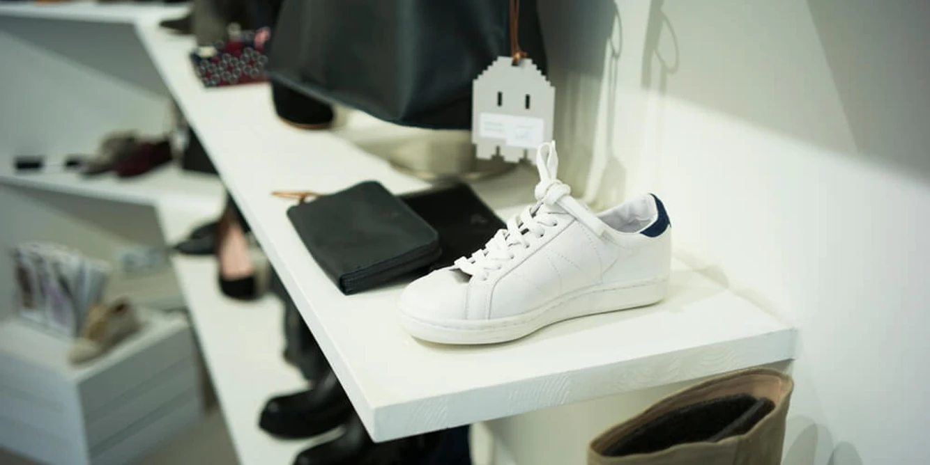 THE WANTS SHOES Weißer Sneaker auf einem Display, umgeben von verschiedenen Accessoires und Schuhen in neutralen Tönen.White sneaker on a display, surrounded by various accessories and shoes in neutral tones.