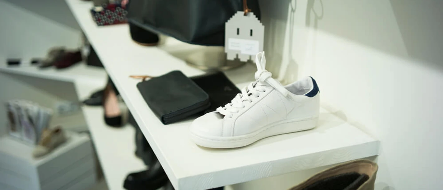 THE WANTS SHOES Weißer Sneaker auf einem Display, umgeben von verschiedenen Accessoires und Schuhen in neutralen Tönen.