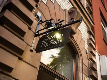 Simon und Renoldi Ein elegantes Schild mit der Aufschrift „Simon und Renoldi“ hängt an einer architektonisch verzierten Hausfassade. An elegant sign reading "Simon und Renoldi" hangs on the façade of an architecturally ornate house.